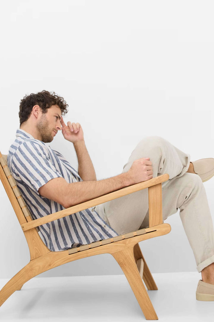 Homme assis sur une chaise en bois, portant une chemise à rayures et un pantalon léger, dans un style décontracté.