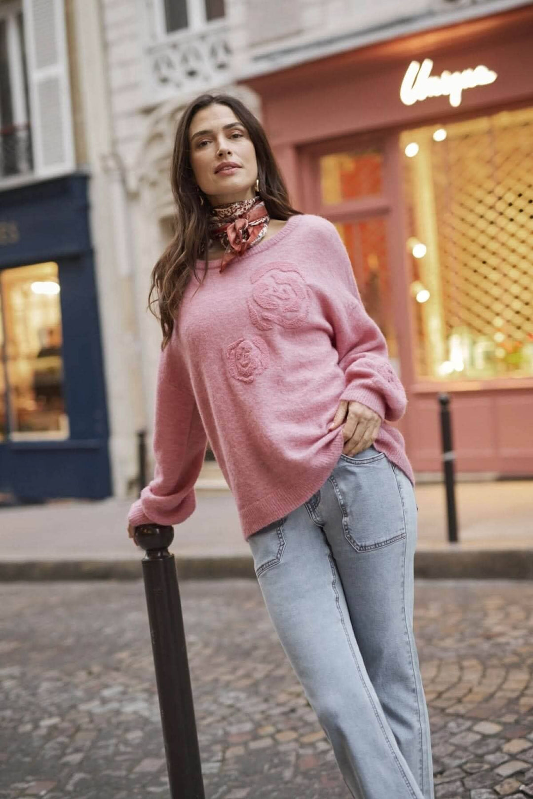 Femme dans un pull rose avec des fleurs, portant un foulard, posant dans une rue parisienne.