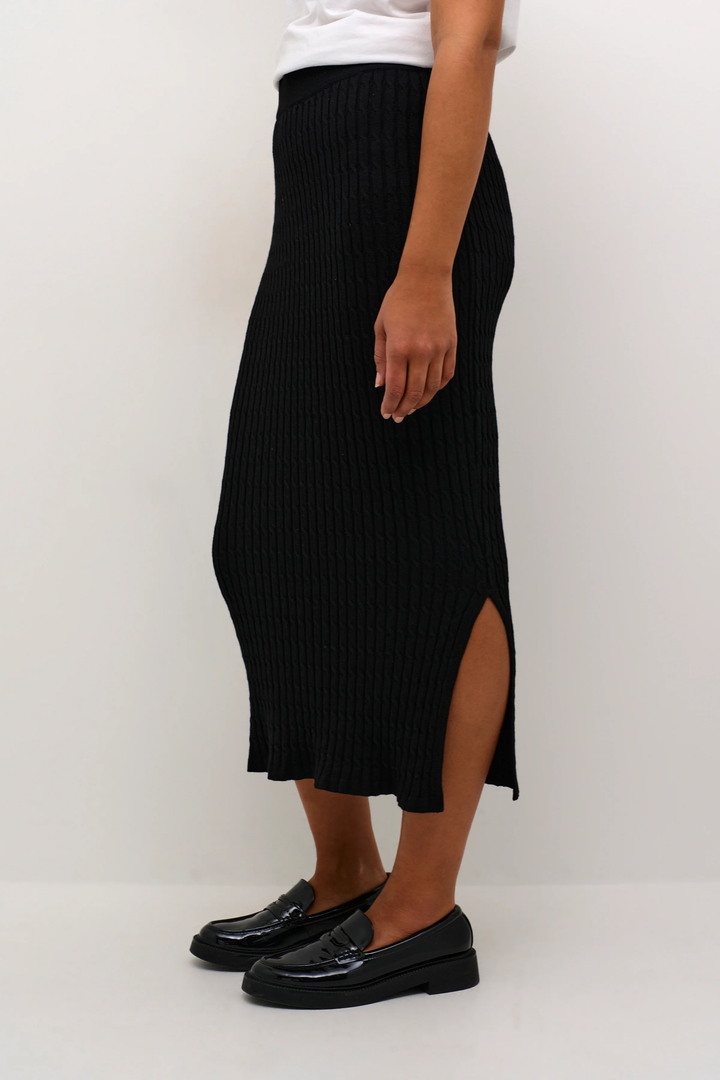 Jupe longue noire ajustée avec fente sur le côté, portée avec des chaussures noires élégantes.