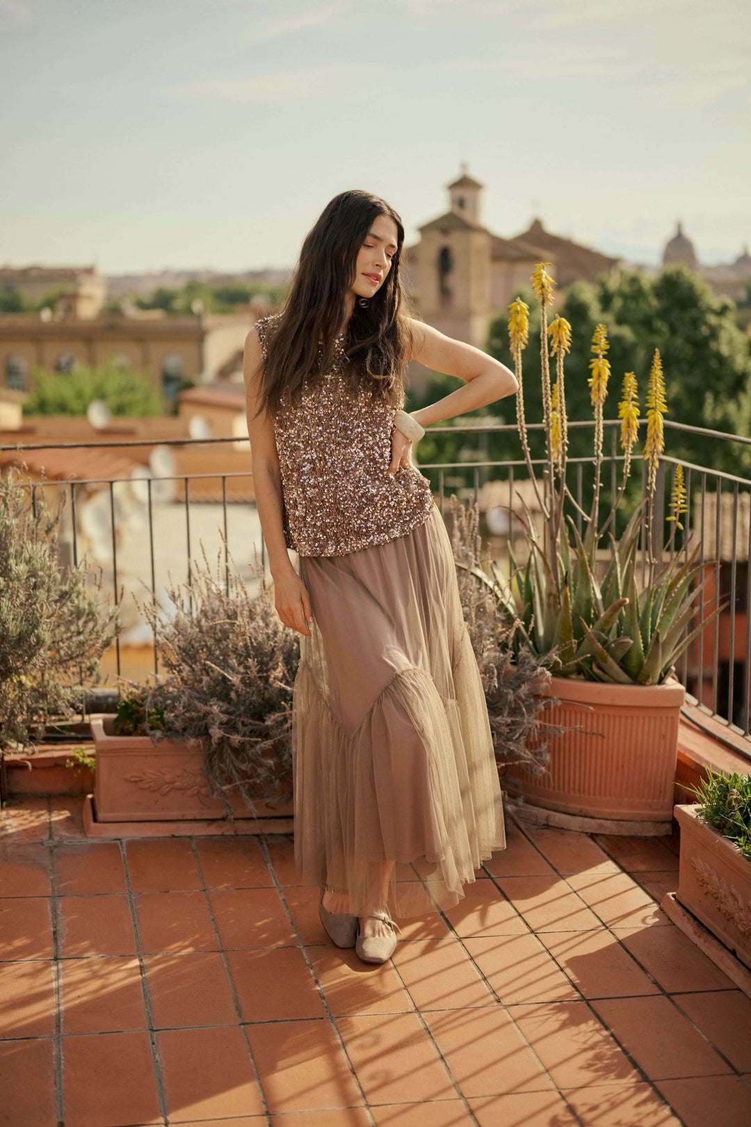 Femme portant une jupe midi en tulle vaporeux sur un balcon ensoleillé, avec des plantes en pot derrière.