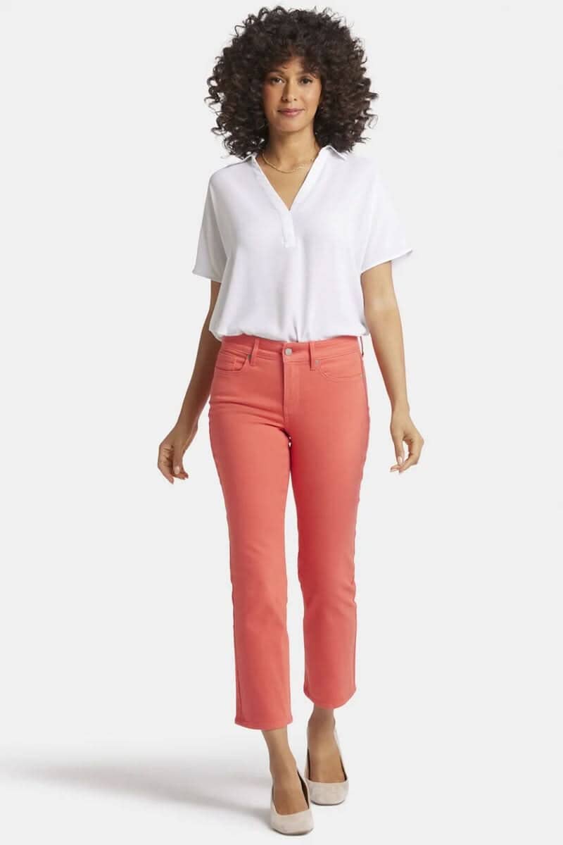 Femme portant un pantalon Marilyn rose corail et un haut blanc, avec cheveux bouclés, debout devant un fond blanc.