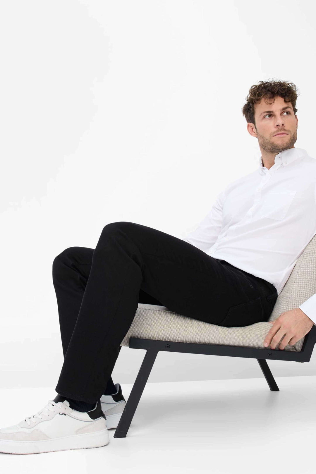Homme assis sur un fauteuil, portant un pantalon noir et une chemise blanche, style moderne et décontracté.