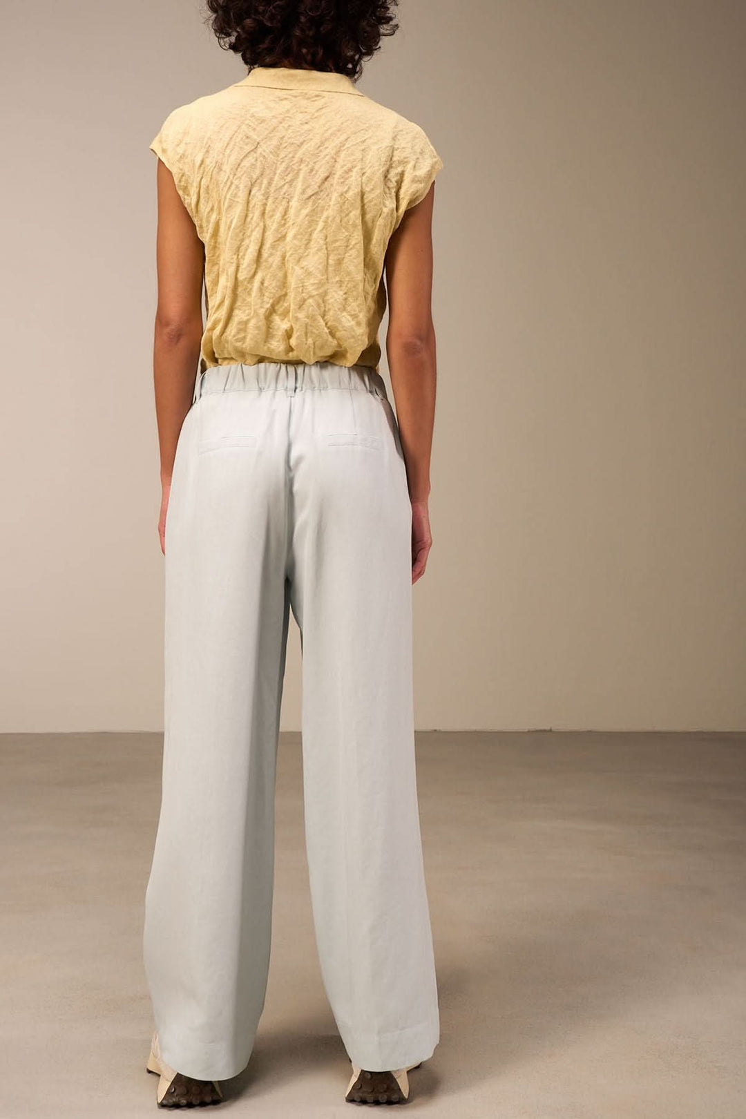 Femme vue de dos portant un pantalon large bleu clair et un haut jaune, dans un cadre minimaliste.