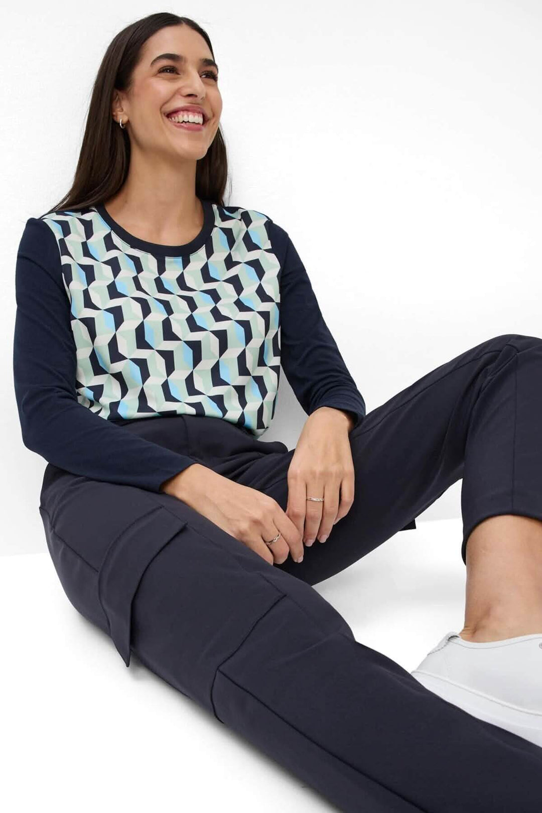 Femme souriante portant un haut graphique et un pantalon ceinturé Morris, assise avec des baskets blanches.