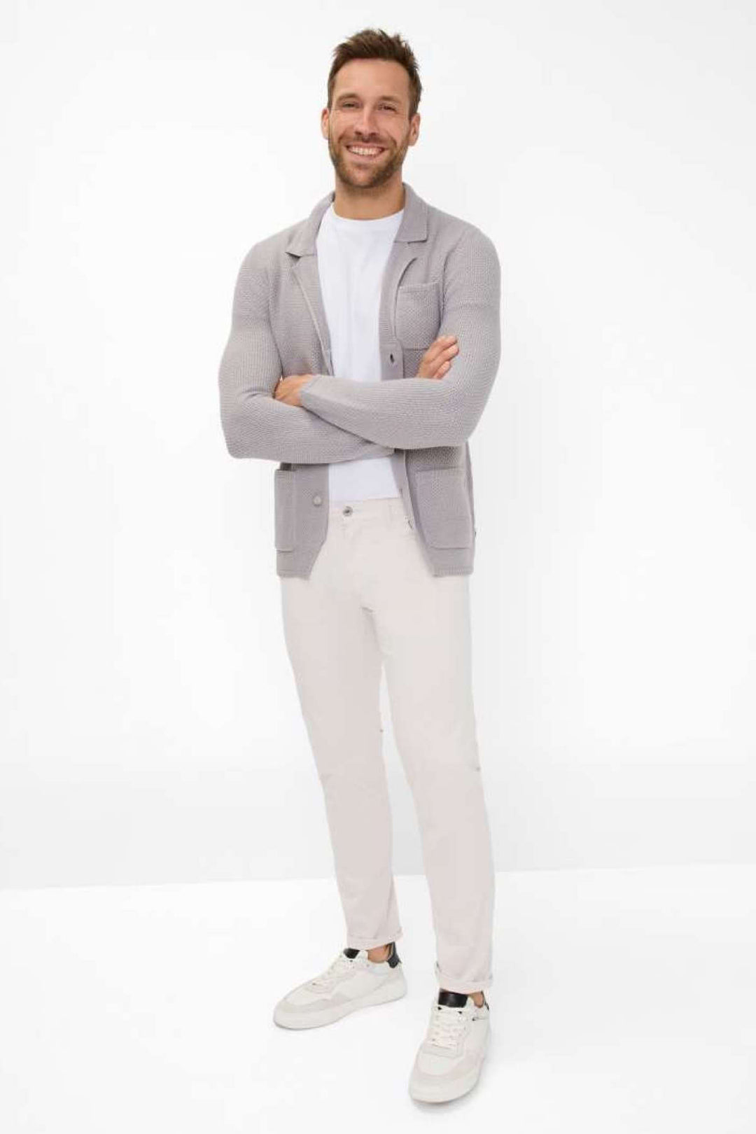 Homme souriant portant un cardigan gris, un t-shirt blanc et un pantalon cinq poches beige, style décontracté chic.