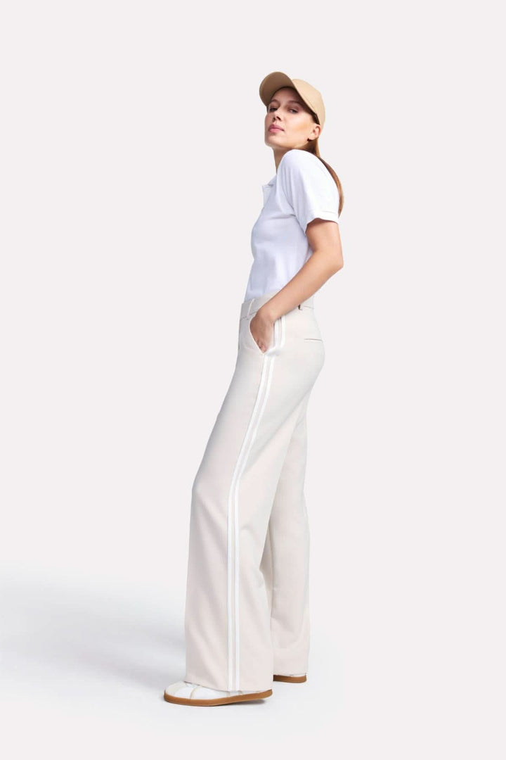 Pantalon classique Amelie à jambe large avec bandes contrastantes, associé à un polo blanc et une casquette beige.