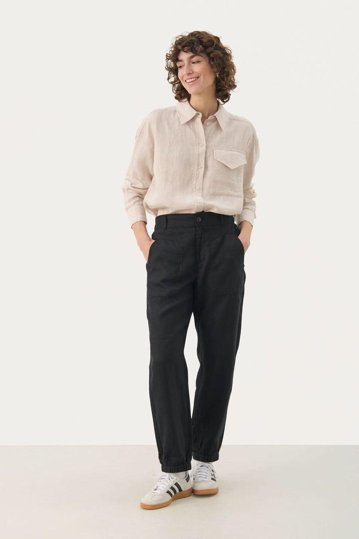 Femme souriante portant un pantalon ample en lin noir avec une chemise beige et des baskets blanches.