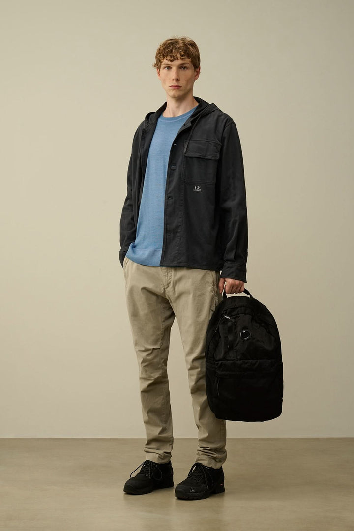 Homme portant un pantalon beige et un hoodie noir, tenant un sac à dos noir, dans un environnement neutre.