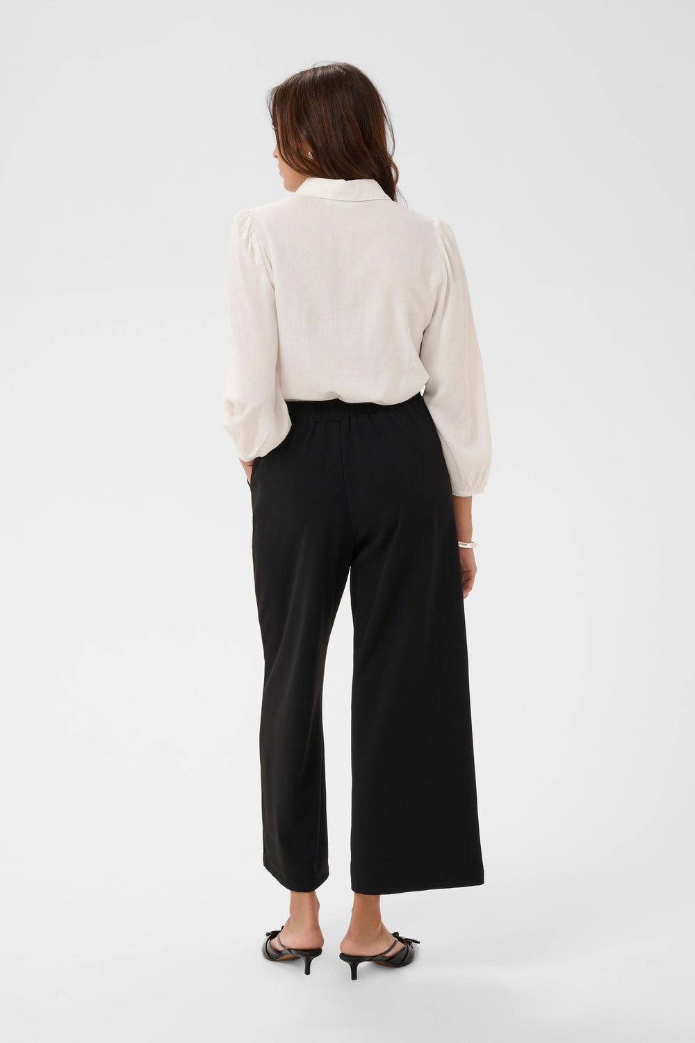Dos d'une femme portant le Pantalon jambe large Phoebe noir, coupe fluide avec ceinture, blouse blanche et talons, style élégant.