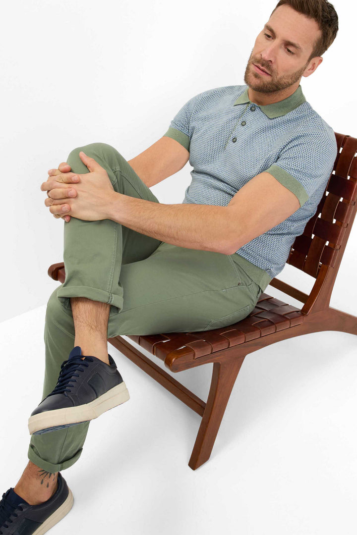 Homme assis sur une chaise en bois, portant un polo bleu et un pantalon marathon vert, avec des sneakers élégantes.