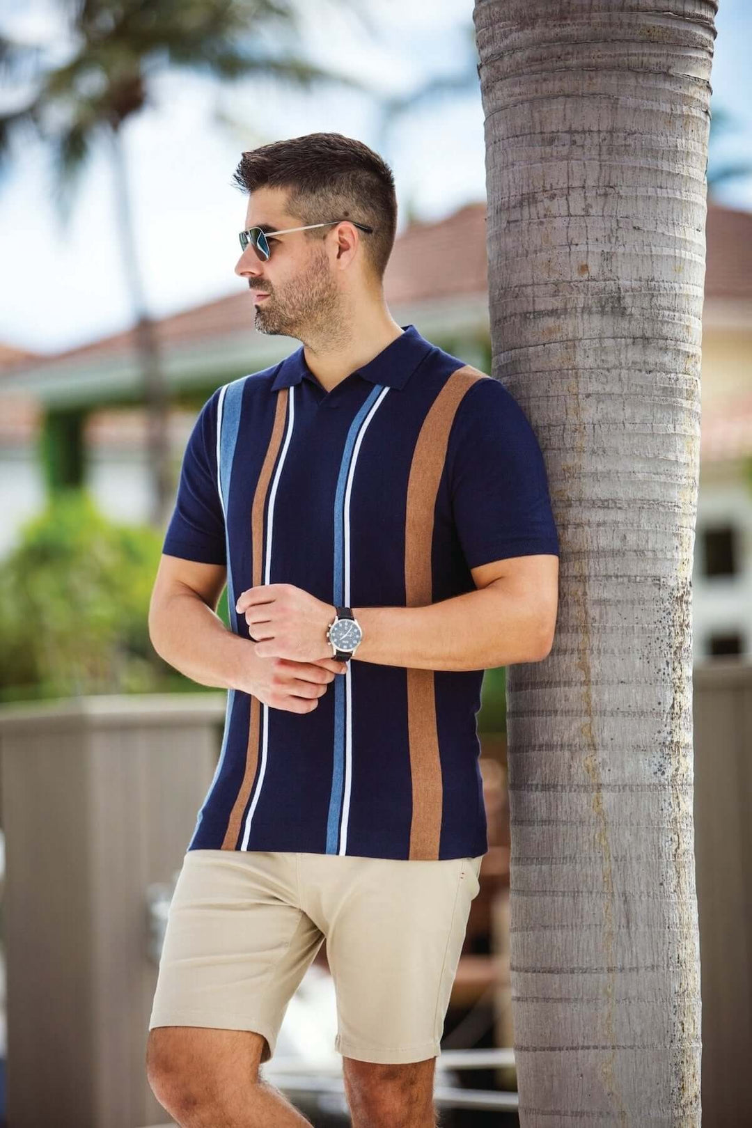 Homme portant un polo à rayures verticales bleues et brunes, accoudé contre un palmier.