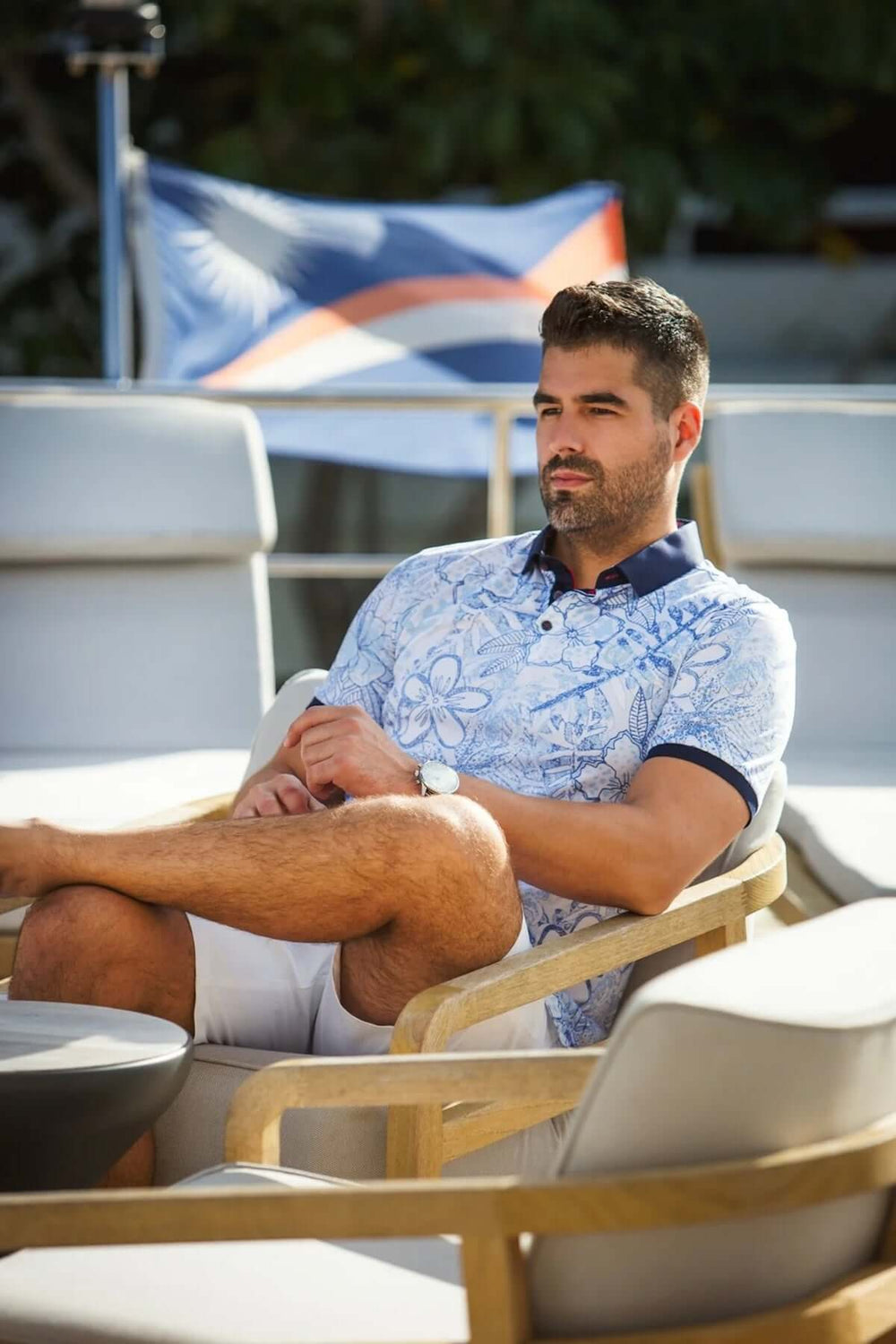 Homme portant un polo à motifs floraux, assis confortablement sur une chaise, décor extérieur élégant.