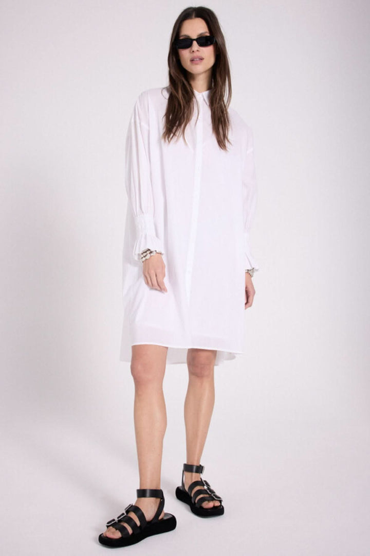 Robe chemise blanche avec détails plissés et cœur brodé, portée avec des sandales noires.