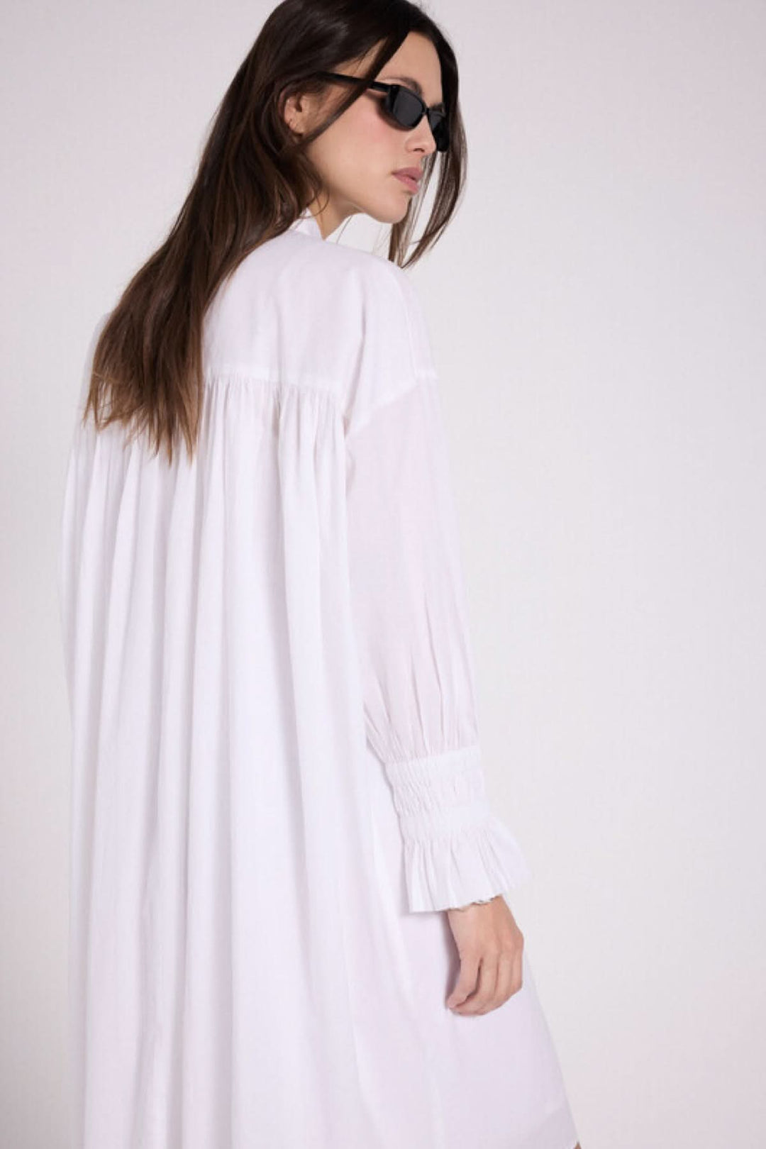 Robe chemise blanche avec détails plissés au dos et manches bouffantes, portée avec des lunettes de soleil.