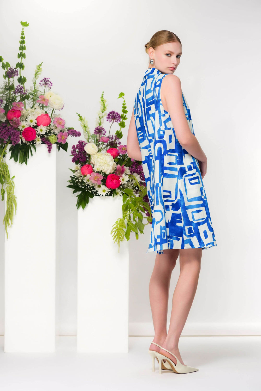 Robe col chemisier sans manches avec imprimé graphique en bleu, portée près de colonnes de fleurs colorées, idéale pour un look estival.