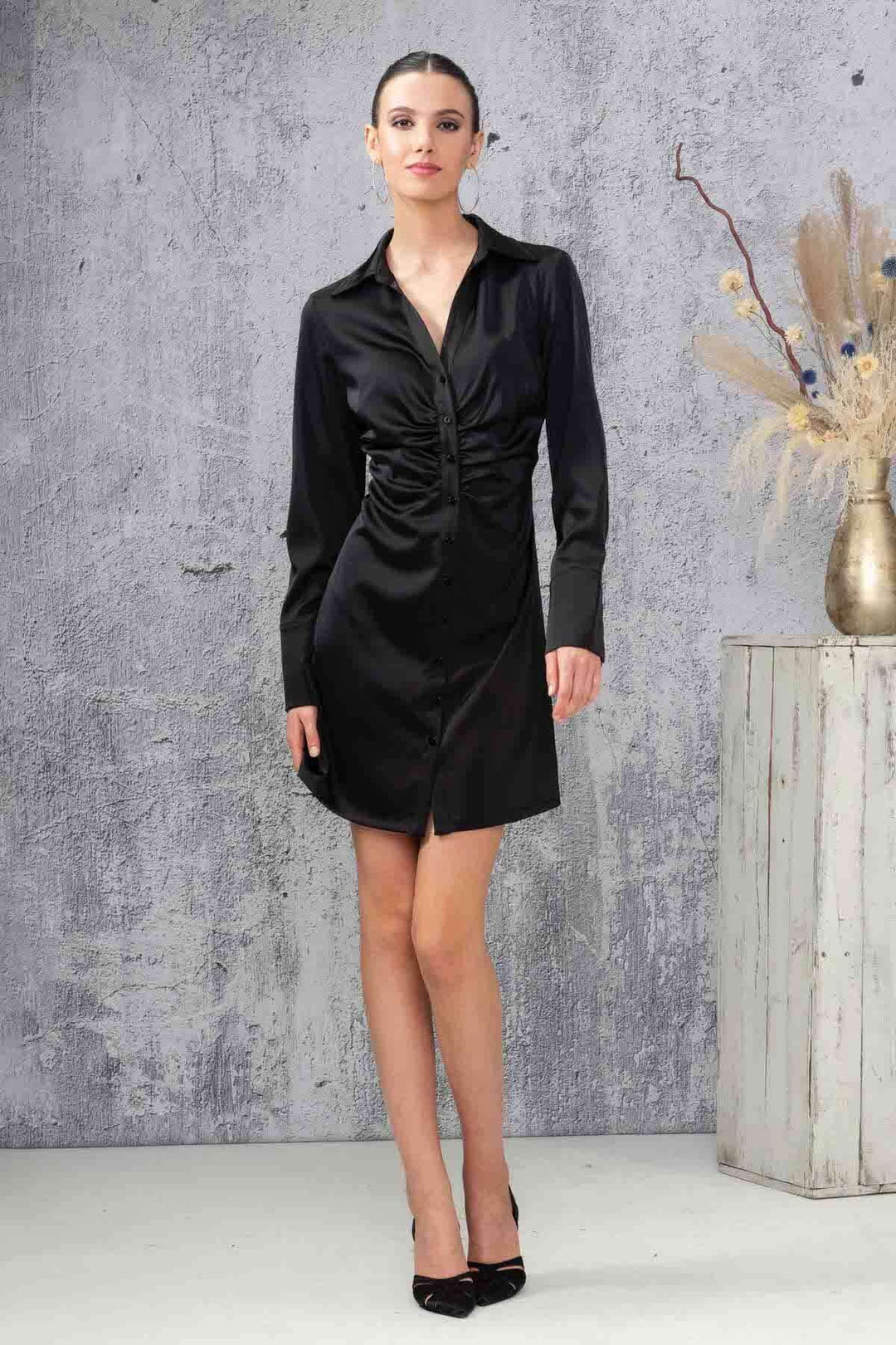 Robe courte noire à manches longues avec col en V et fronces, portée par une femme élégante, sur fond texturé.