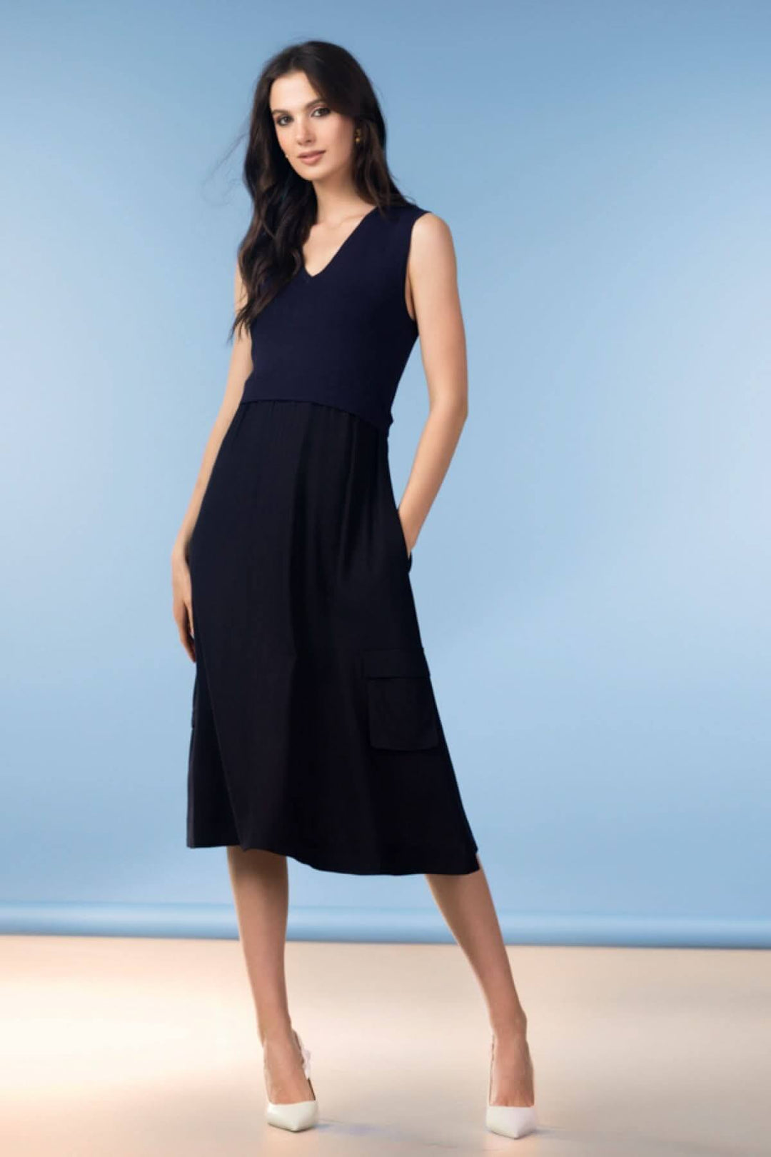 Femme portant une robe midi fluide col en V, sans manches, couleur sombre, avec poches plaquées et coupe moderne sur fond bleu.