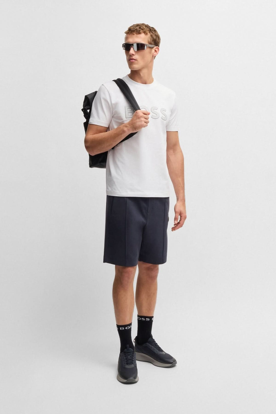 Homme portant un t-shirt blanc avec logo BOSS, short noir, lunettes de soleil et sac à dos.