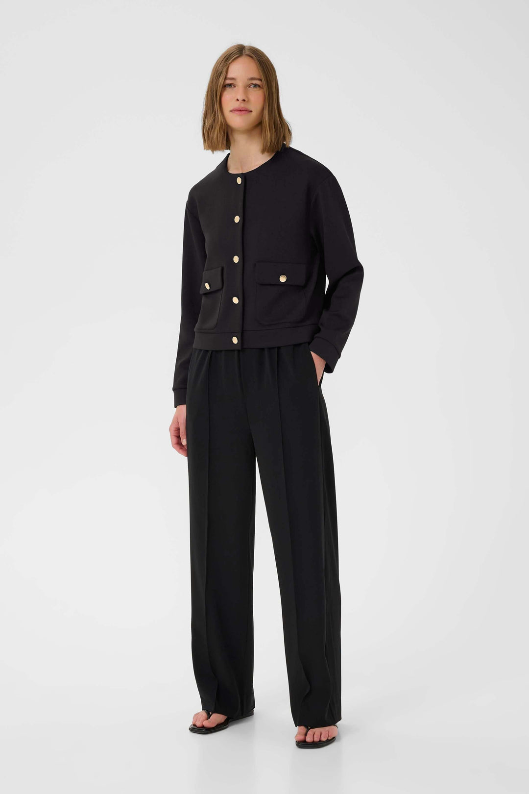 Veste Gida noire à col rond avec boutons dorés, portée avec un pantalon noir, style élégant et moderne.