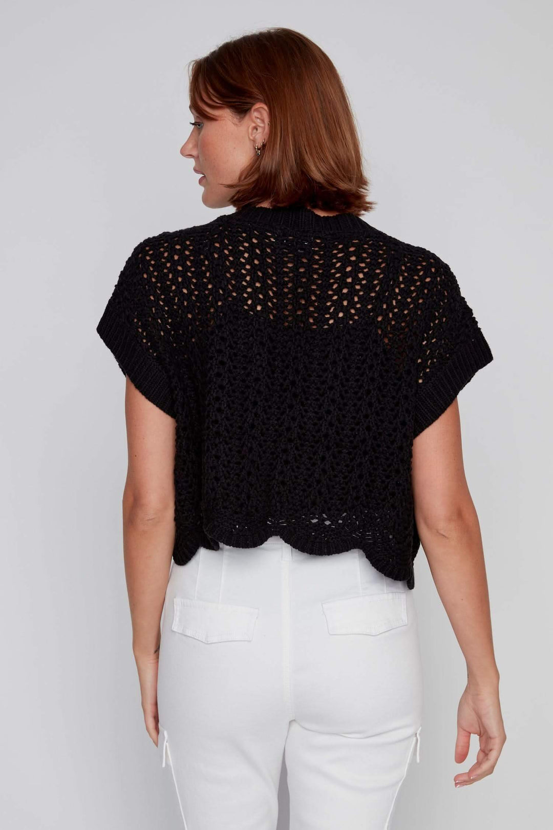 Veste au crochet noire, à manches courtes, à tricot ajouré, portée avec un pantalon blanc.