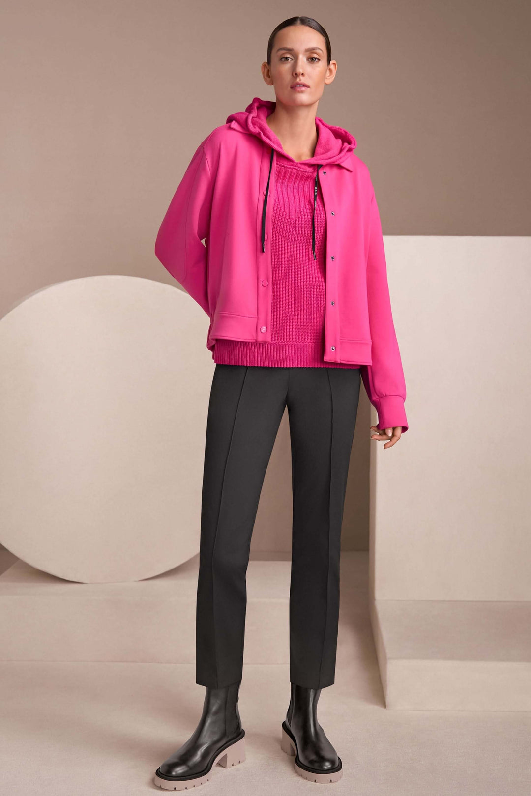 Femme en veste rose moderne avec pull à col montant et pantalon noir, pose décontractée sur fond neutre.