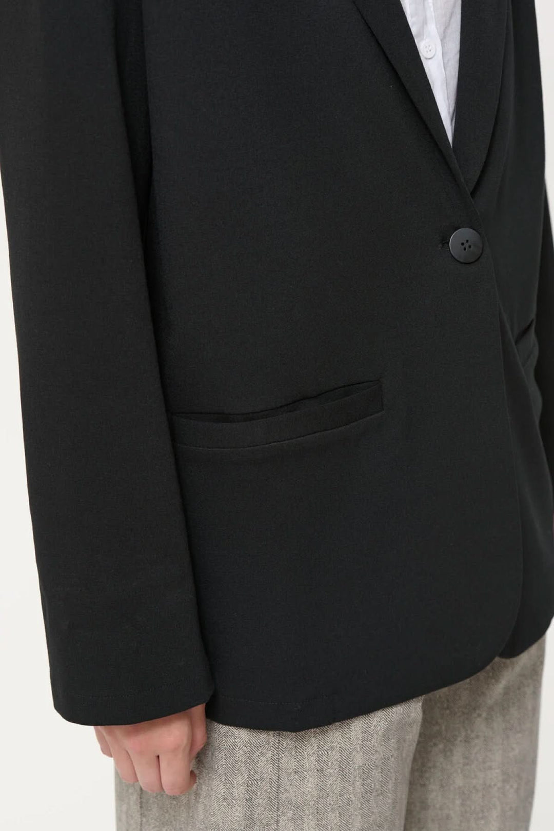 Détail d'un veston Cayden noir, à la coupe ample, avec fausses poches et manche longue, porté sur un pantalon beige.