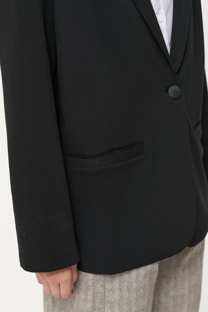 Détail d'un veston Cayden noir, à la coupe ample, avec fausses poches et manche longue, porté sur un pantalon beige.