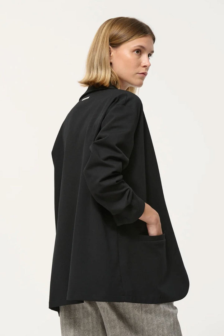 Veston Cayden noir, coupe droite ample, manches longues, fausses poches passepoilées, porté ouvert sur tenue ajustée.