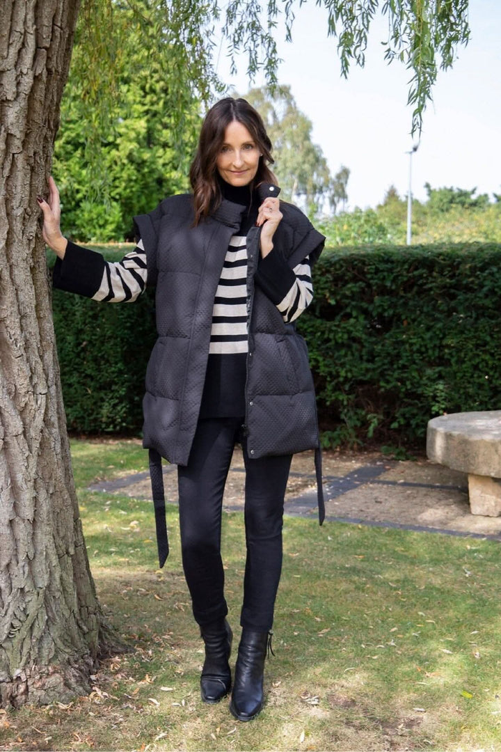 Femme souriante portant un gilet matelassé noir Tahani, debout près d'un arbre en plein air.