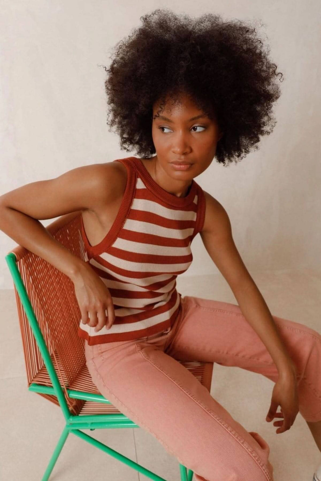 Femme portant un pull à bretelles rayé marron et crème, assise sur une chaise colorée.