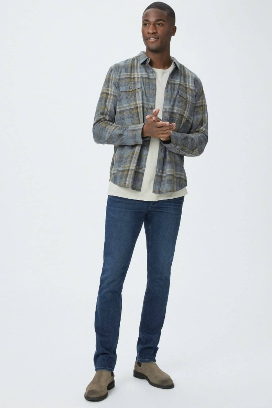 Homme souriant portant une chemise à carreaux bleue et un t-shirt beige, avec un jean slim bleu foncé et des bottes beige.