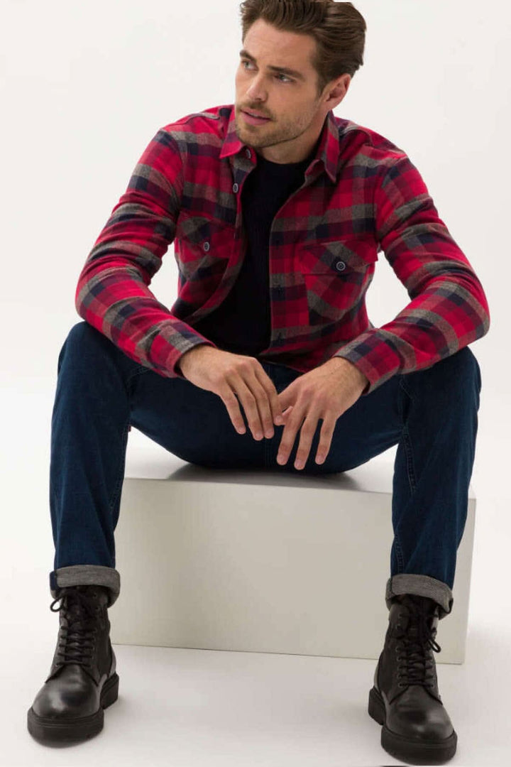 Homme assis portant une chemise à carreaux rouge et noir, avec jeans modernes et chaussures noires, sur une plateforme blanche.