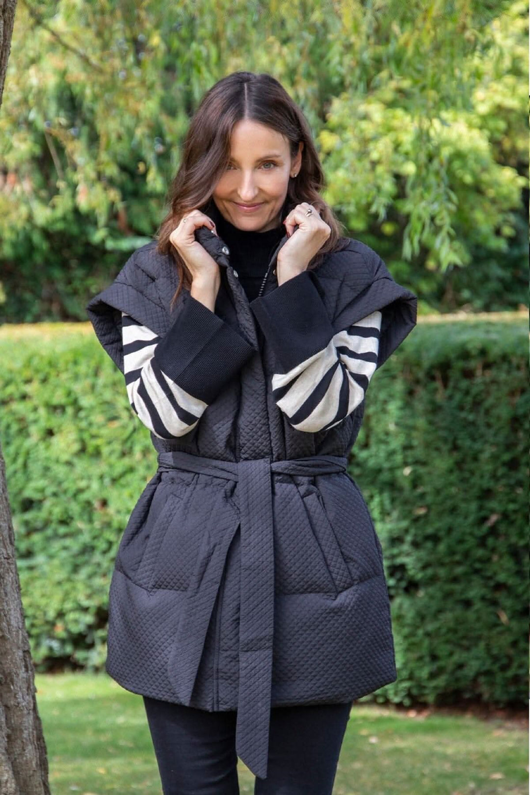 Femme portant un gilet matelassé noir Tahani avec ceinture, posé dans un jardin.