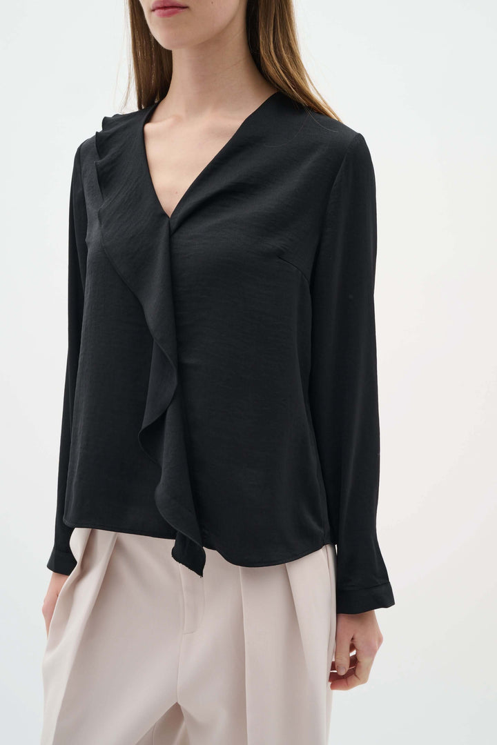 Blouse Auda noire avec col en V et détail volanté, porté avec un pantalon clair pour un look chic.