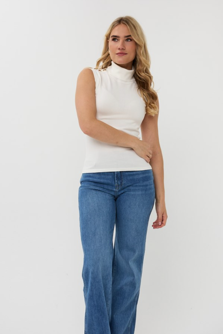 Col roulé sans manches blanc avec boutons métalliques, porté avec un jean bleu pour un look chic et élégant.