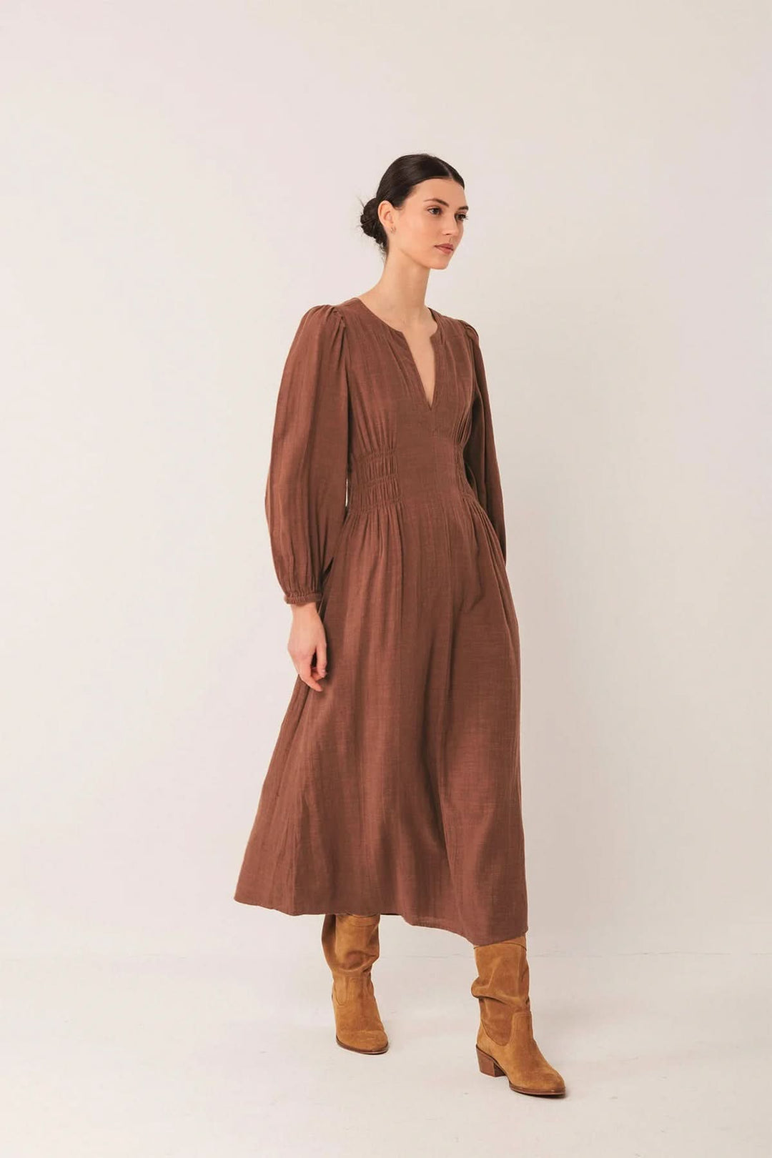 Femme portant une robe longue à encolure V et manches bouffantes avec bottes, silhouette moderne et élégante. Décris moi le produit en français.