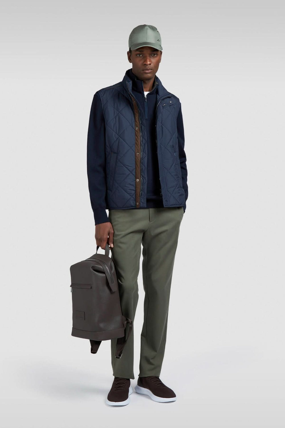 Homme portant un gilet matelassé bleu, pantalon vert, et casquette, tenant un sac à dos élégant.