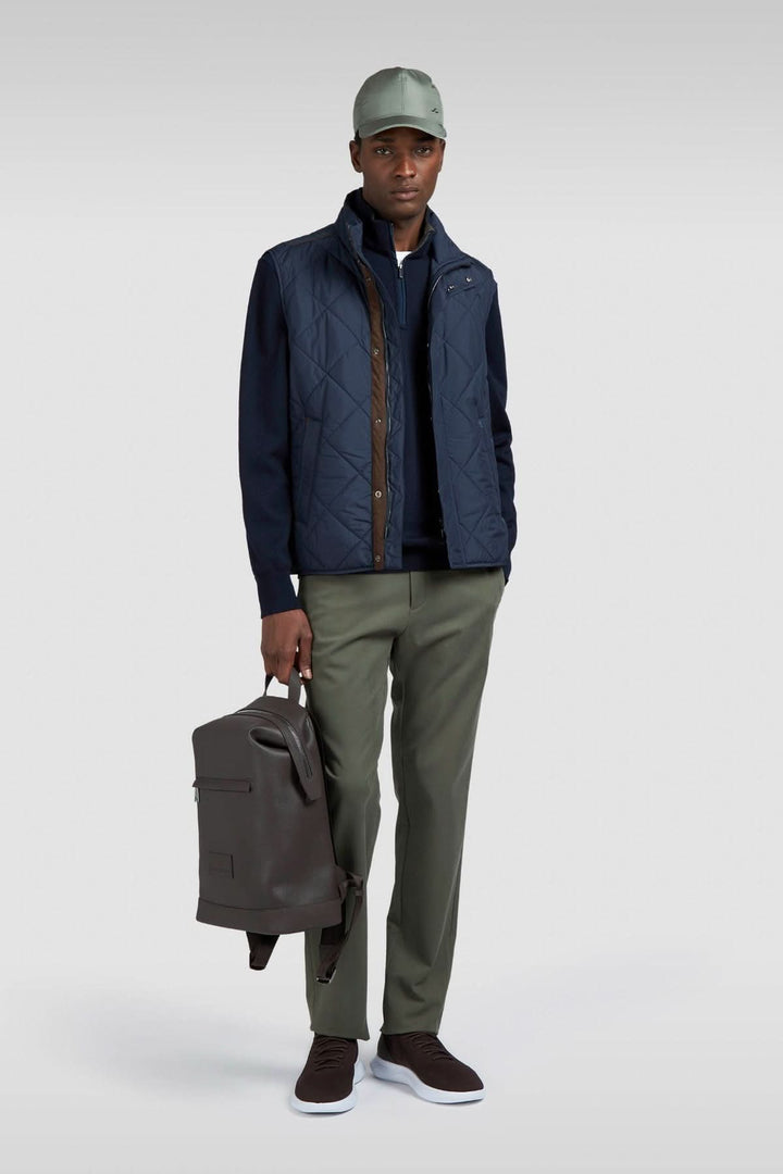 Homme portant un gilet matelassé bleu, pantalon vert, et casquette, tenant un sac à dos élégant.