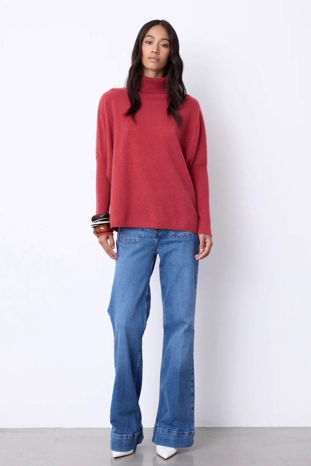 Femme portant un chandail à col roulé rouge en cachemire, élégant et confortable, associé à un jean droit et des bracelets, pour une allure chic.