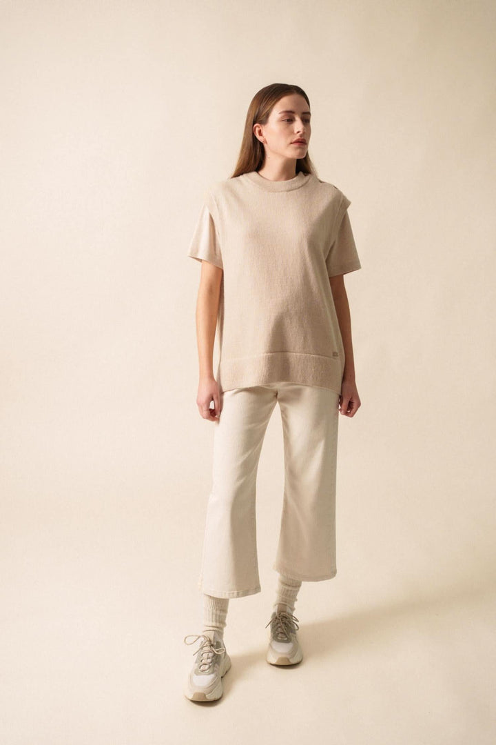 Femme portant un t-shirt beige et un pantalon clair, posant sur fond neutre, style décontracté.