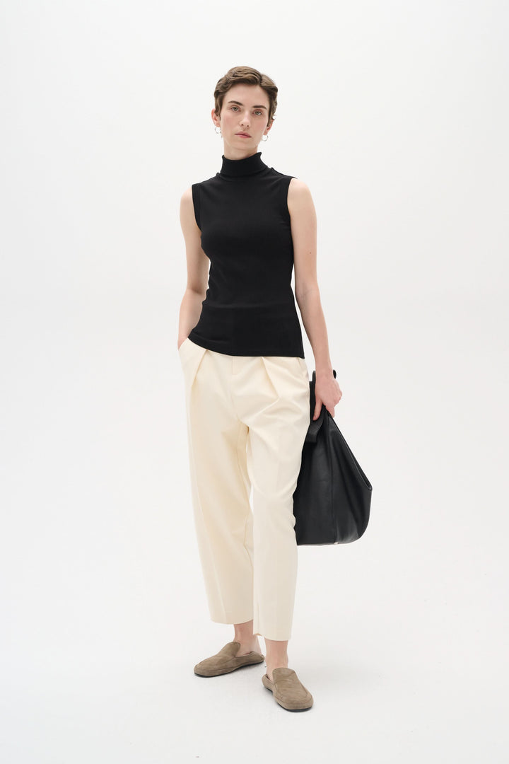 Modèle portant une camisole noire sans manches à col montant, pantalon beige et sac noir, pose décontractée.