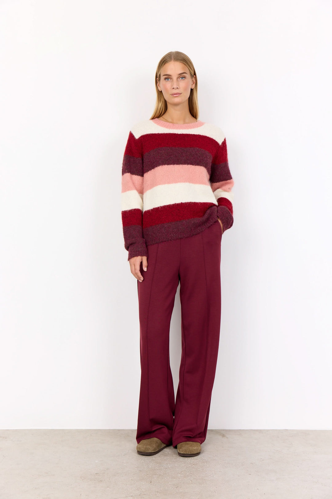 Femme portant un tricot à rayures larges dans des tons de rose et bordeaux, avec un pantalon bordeaux, sur fond blanc.