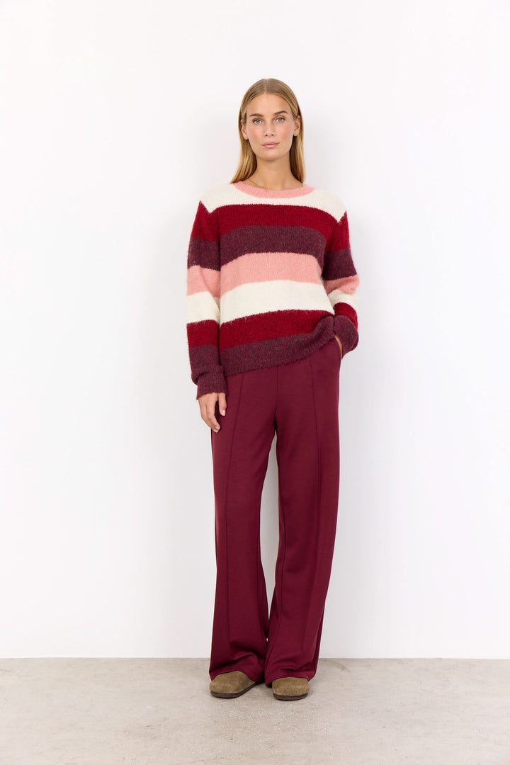 Femme portant un tricot à rayures larges dans des tons de rose et bordeaux, avec un pantalon bordeaux, sur fond blanc.