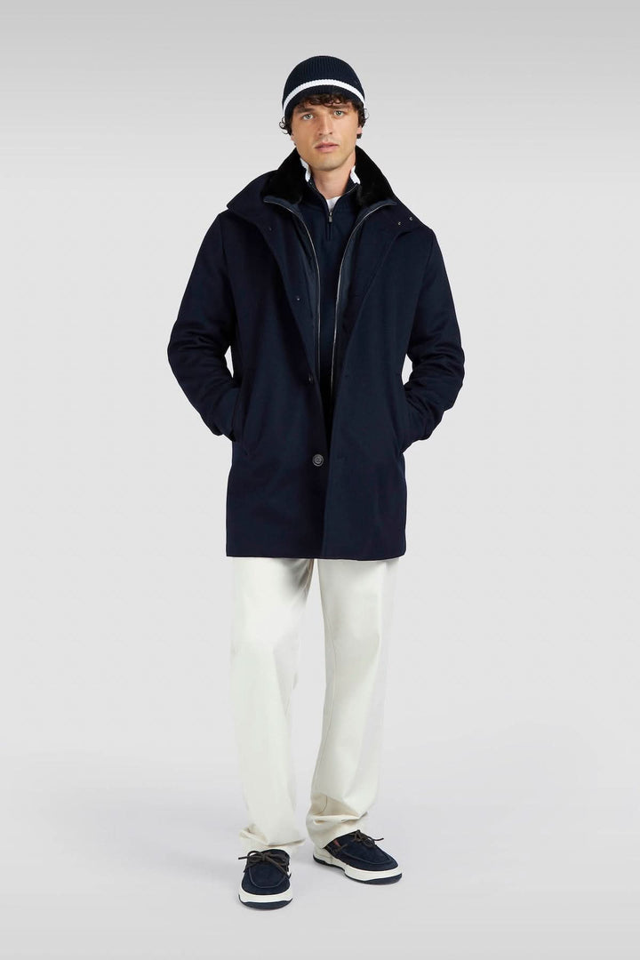 Homme portant un manteau bleu marine avec un chapeau, pantalon clair et chaussures, look décontracté et élégant.