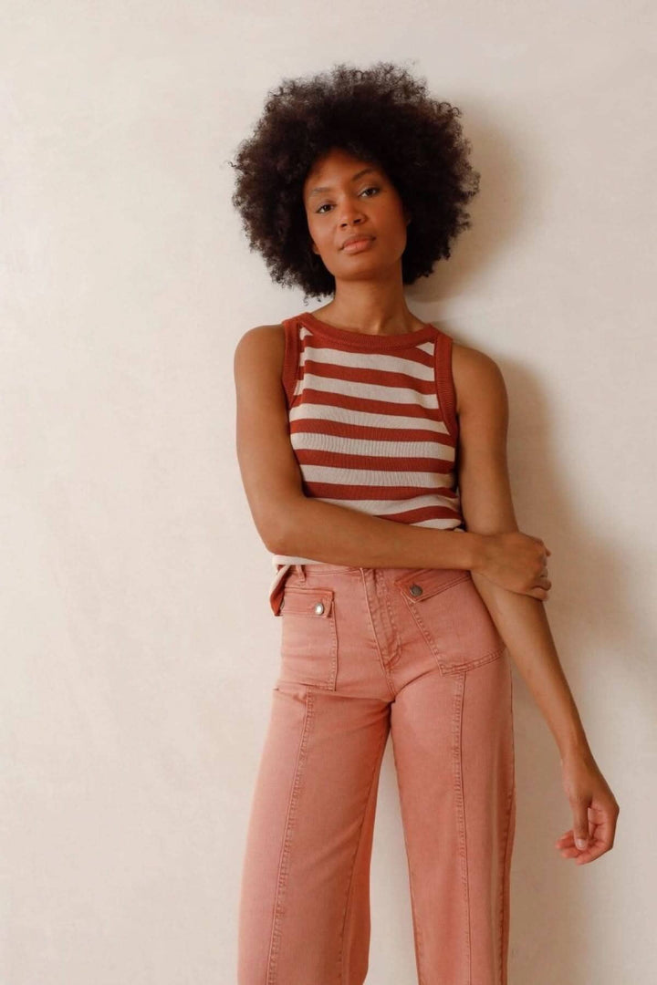 Femme avec un haut à rayures marron et crème, portant un pantalon rose, pose contre un mur neutre.