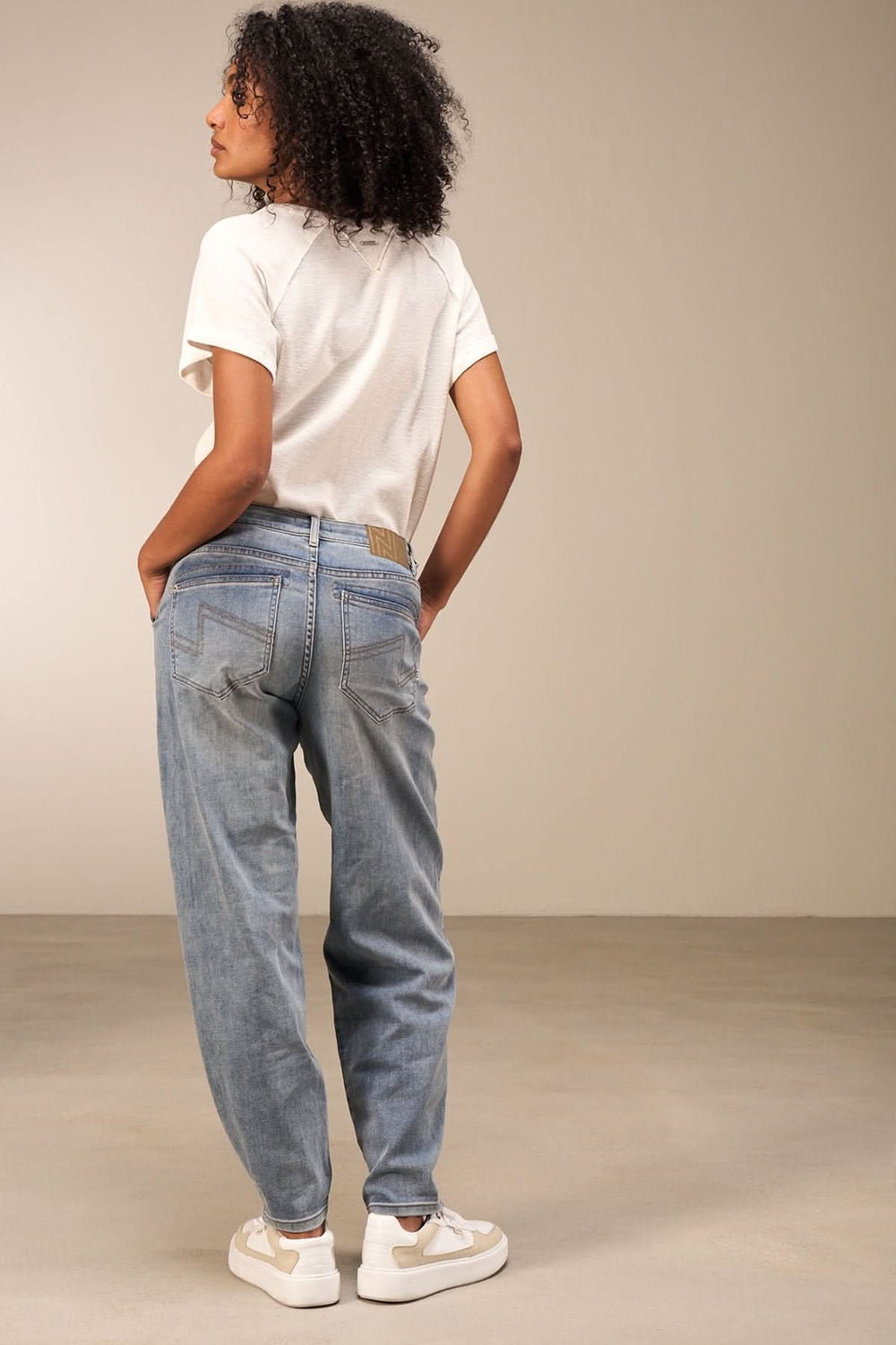 Femme vue de dos portant un jean coupe droite, t-shirt blanc et baskets, dans un cadre minimaliste.
