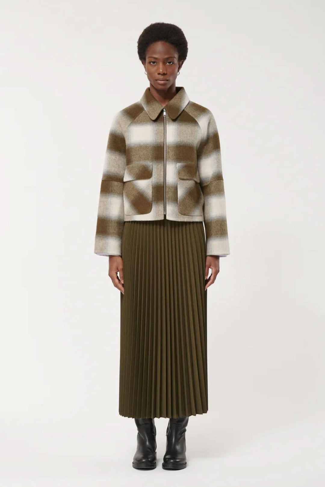 Femme portant une veste courte à carreaux avec poches à rabat et fermeture éclair, associée à une jupe plissée et des bottes noires.