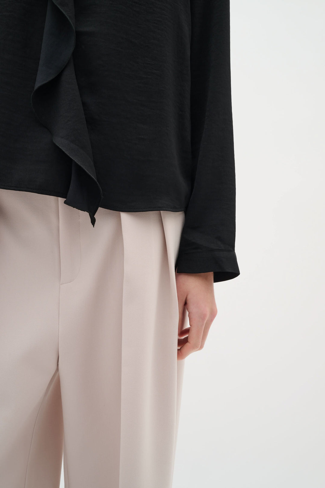 Blouse Auda fluide noire avec détail volanté, portée avec un pantalon à plis beige pour un look élégant.