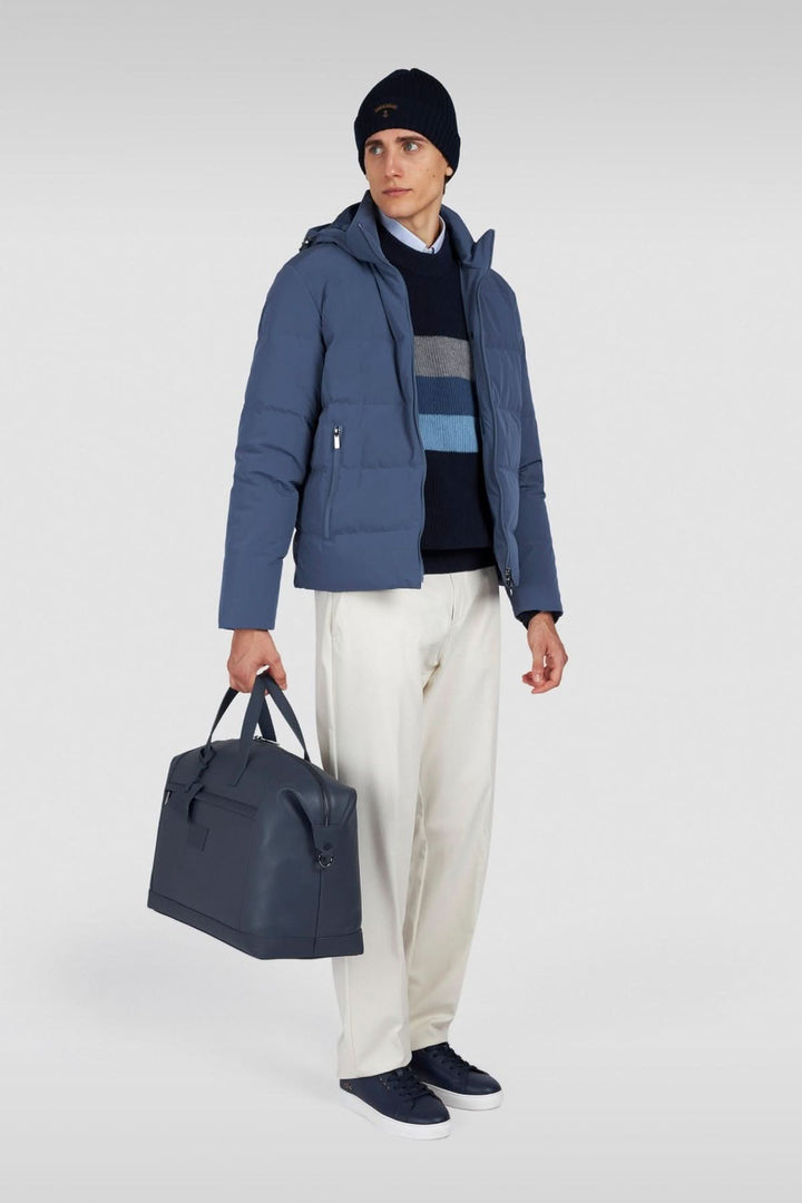 Homme portant un manteau bleu, un pull à rayures, pantalon clair et sac en cuir, créant un look urbain chic.