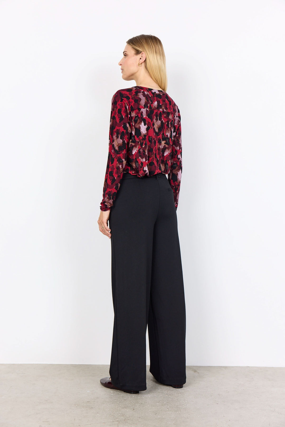 Femme portant une blouse imprimée rouge et noire, vue de dos, avec un pantalon large sur fond blanc.